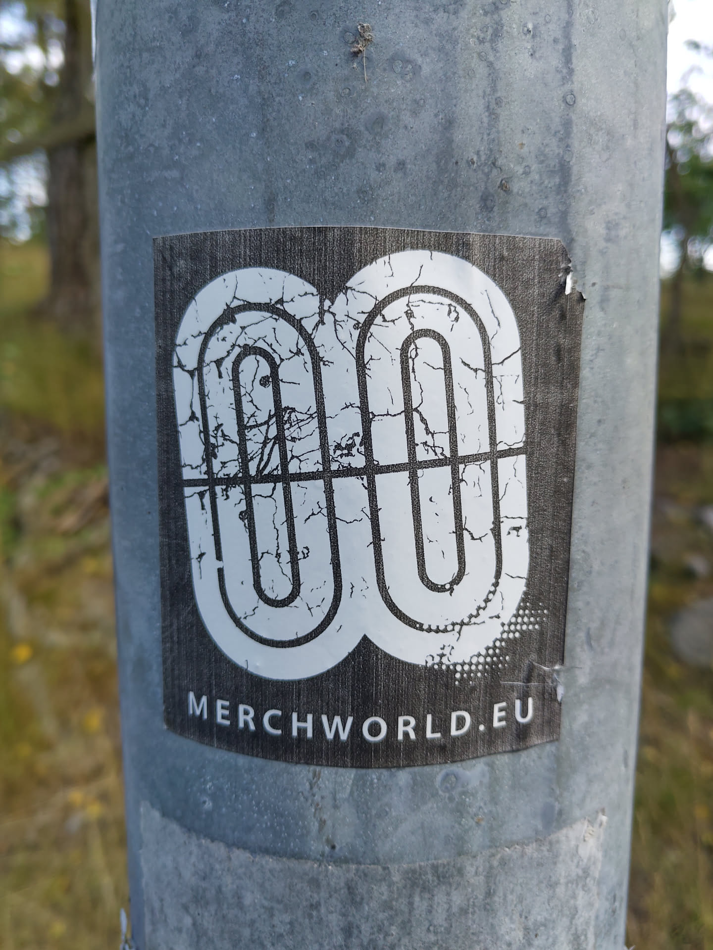 This sticker features a stylized logo resembling interconnected loops, presented in a distressed, cracked style.  The text 'MERCHWORLD.EU' is visible at the bottom.  The sticker appears to be affixed to a metal pole outdoors.