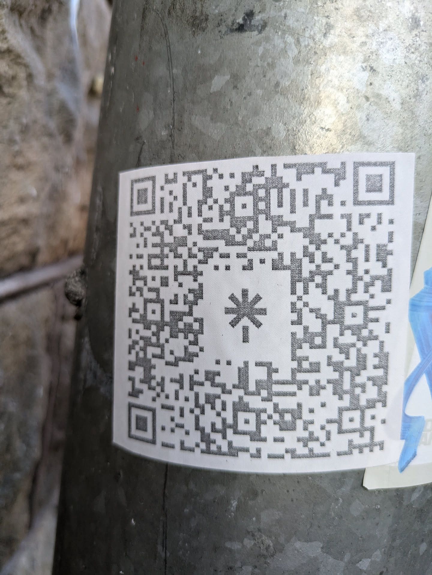 A close-up shot of a square QR code sticker affixed to a weathered metal pole. The QR code is predominantly black and white, with a small asterisk symbol in its center. The sticker appears to be made of standard paper and shows slight creases from adhering to a curved surface.