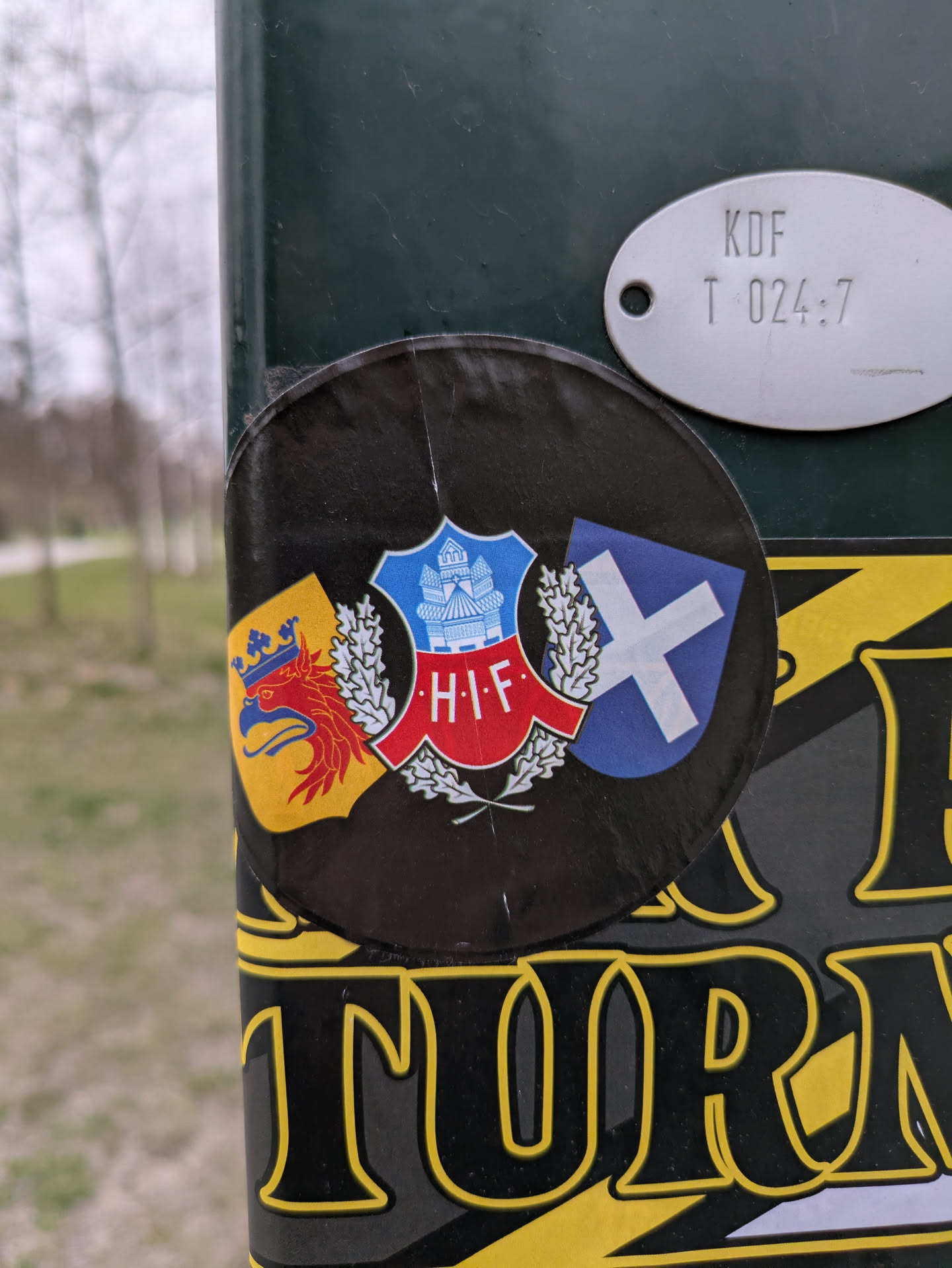 The sticker shows three shields, each with different designs and colors. The central shield features a blue and white design with a crown and buildings, surrounded by laurel. To the left is a shield with a red griffin on a yellow background, and to the right, a blue shield with a white saltire. The letters 'H.I.F.' are prominently displayed on the central shield.