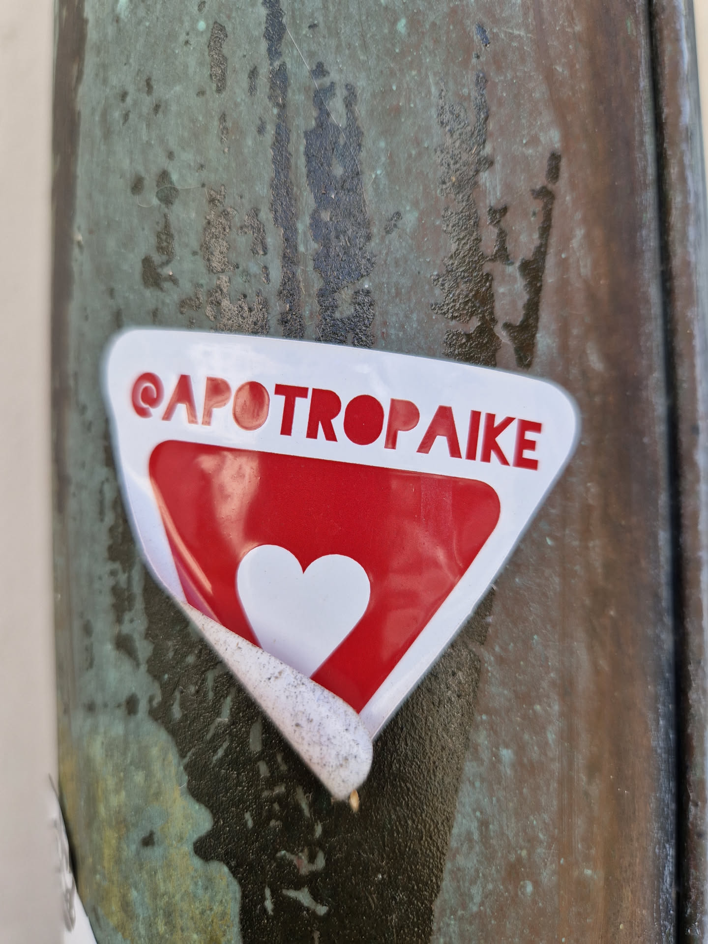A red triangular sticker with a white heart in the center. The word '@APOTROPAIKE' is written at the top. The sticker is slightly peeled off from the surface.