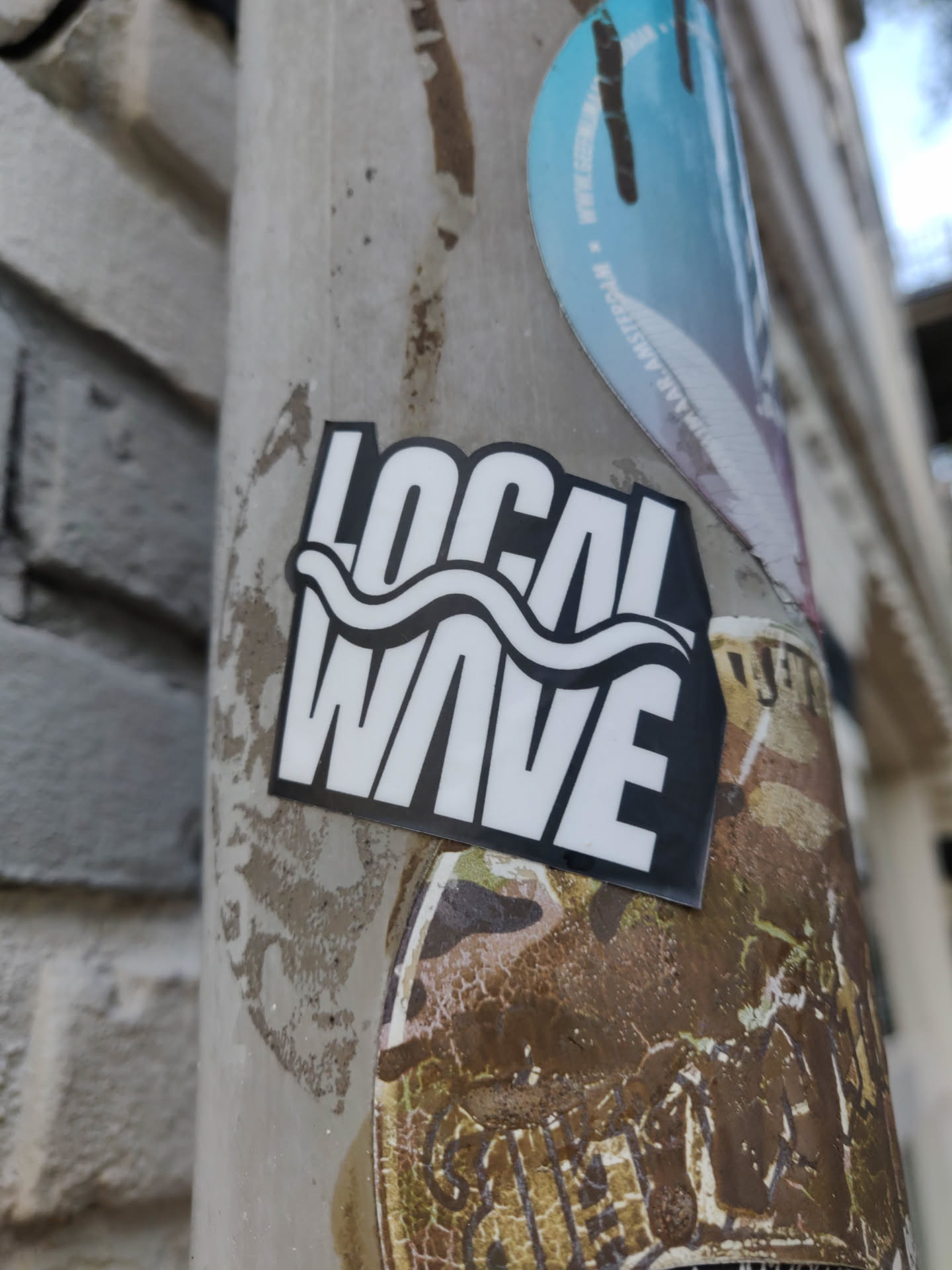 A sticker with the text "LOCAL WAVE" in bold white letters on a black background. The word WAVE includes a simple wave design. It's stuck on a weathered pole with other stickers.