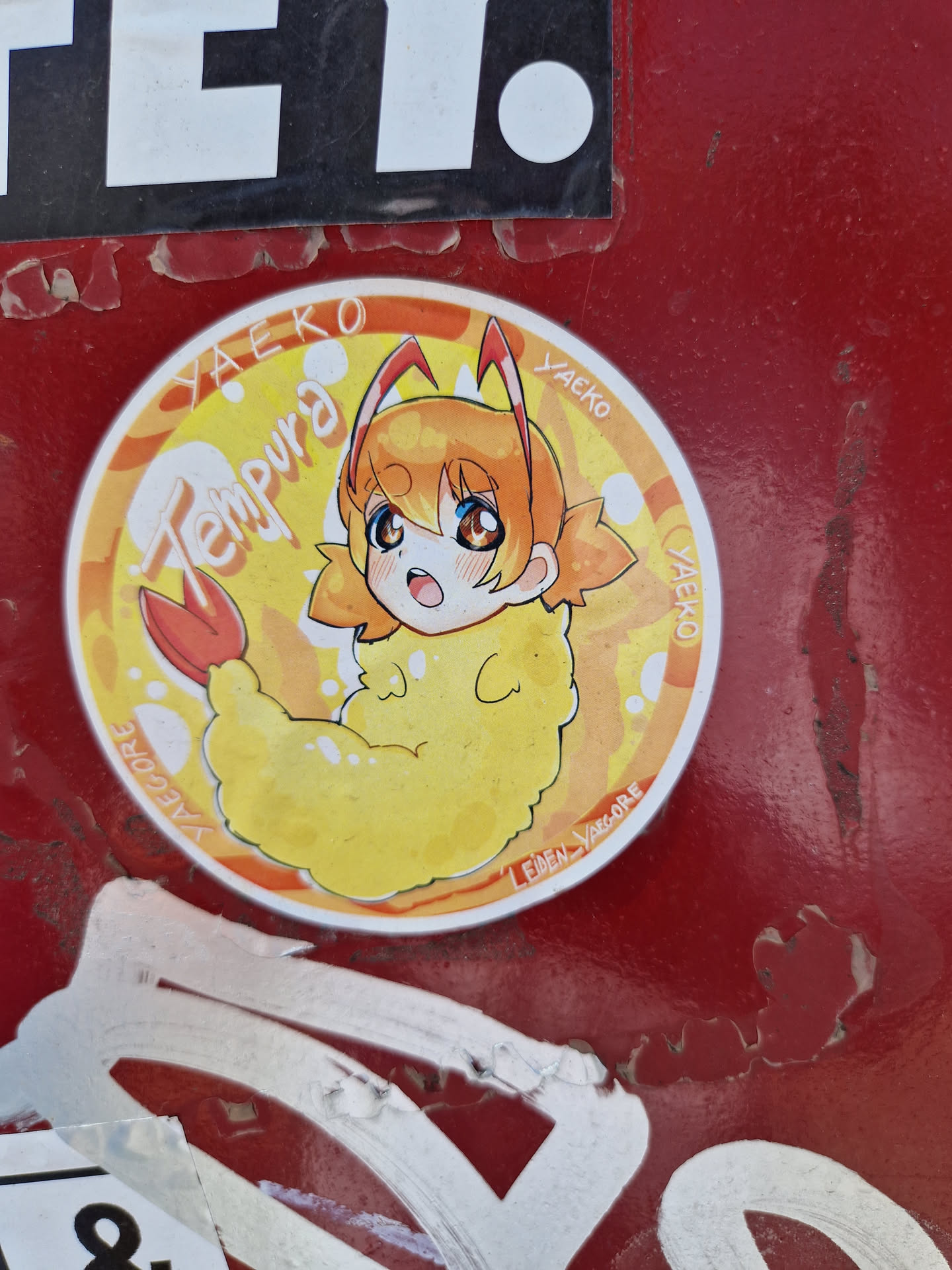 Round sticker featuring an illustration of a cute anime-style girl dressed as a shrimp tempura. The character has orange hair and is drawn in a chibi style. The background is yellow with orange accents. The artist's signature is visible on the sticker.