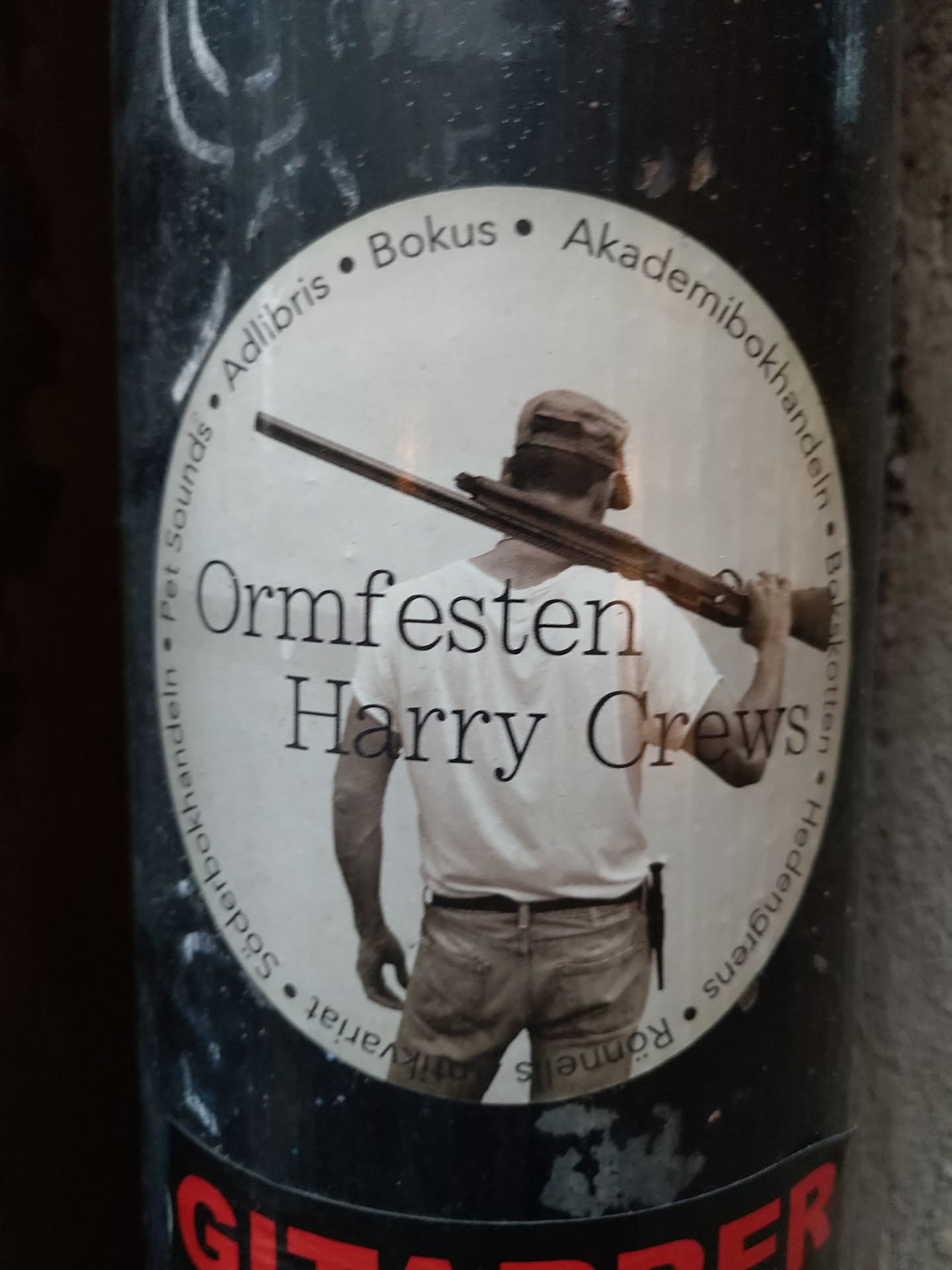 A round sticker featuring a man with a gun on his shoulder. The text on the sticker mentions the book "Ormfesten" by Harry Crews and several bookstores.