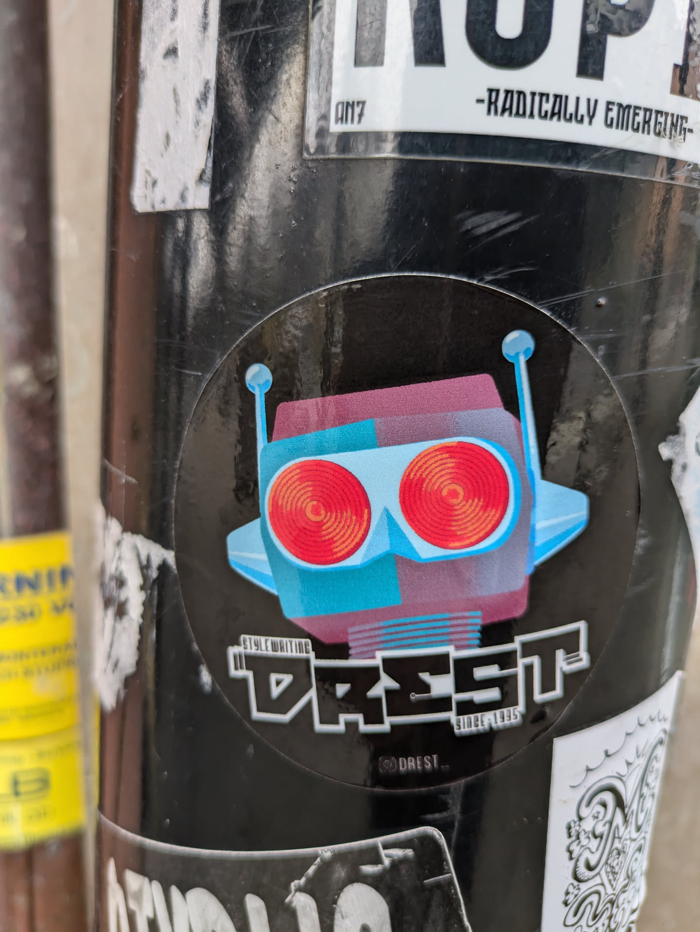 A round sticker featuring a stylized robot illustration. The robot is primarily teal and purple, with red circular accents resembling eyes. The text 'DREST.' is prominently displayed beneath the robot, accompanied by the tagline 'Since 1995'. The sticker has a black background, which gives a nice contrast to the illustration and text.