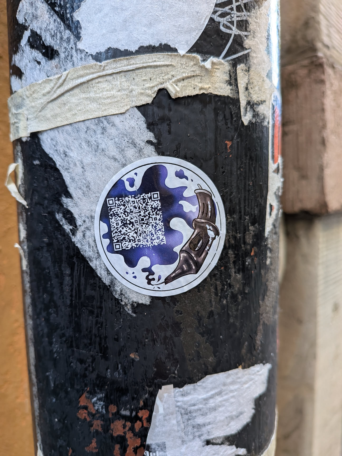 A round sticker adhered to a worn black pole. The sticker features an artistic design comprising an antique-style fountain pen nib on the right edge, pointing downwards and releasing a stylized, splatter of deep purple and white ink. Integrated inside the purple ink puddle on the left is a square QR code.