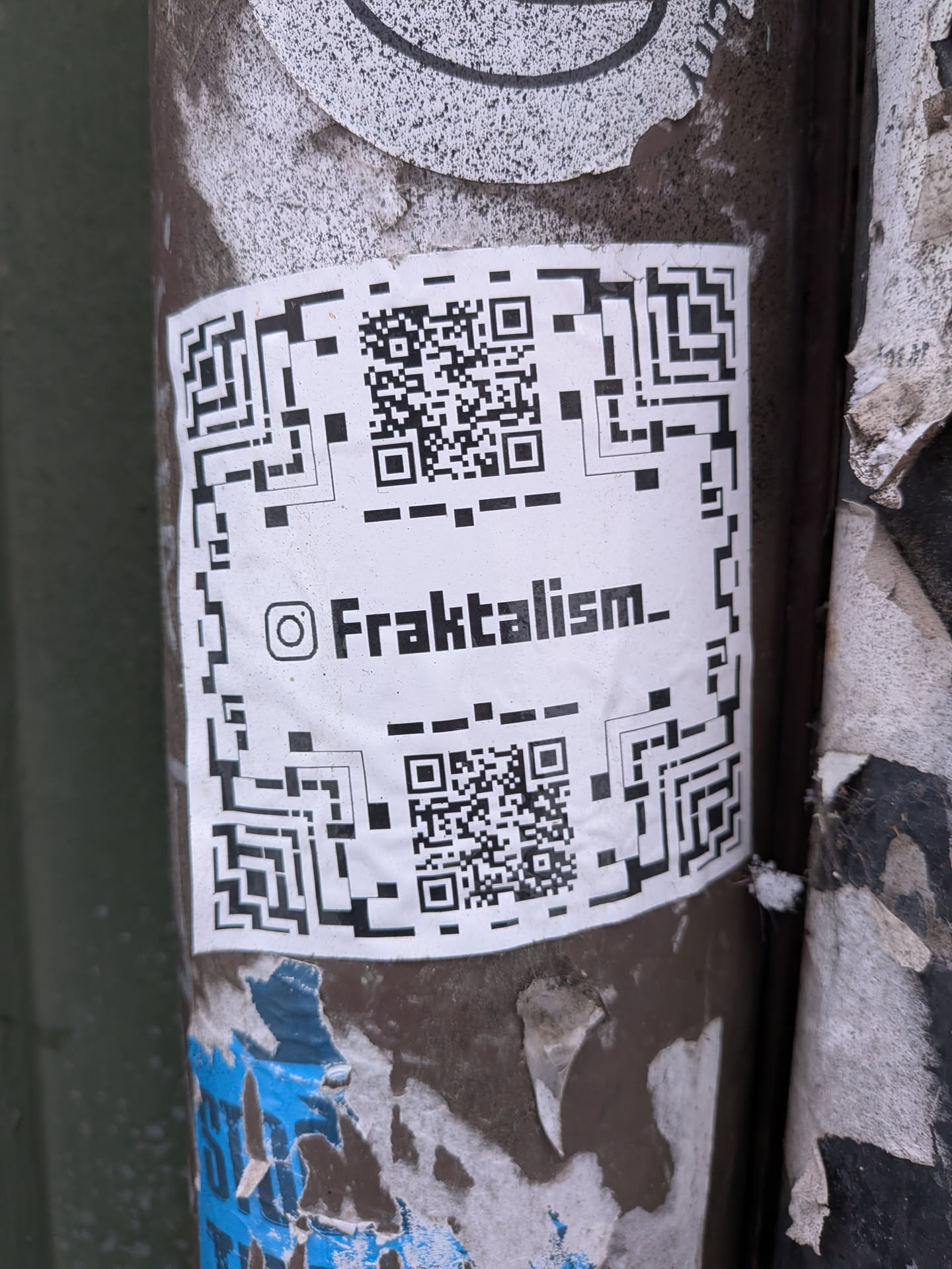 A rectangular white sticker adhered to a dark, textured pole, featuring two prominent black and white QR codes—one at the top and one at the bottom. Centered between the QR codes is the Instagram logo followed by the text 'Fraktalism_'. The sticker shows signs of wear, with slight creases and dirt, indicating its outdoor placement. It is surrounded by remnants of other torn and faded papers.
