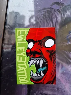 A sticker featuring a demonic red face with sharp teeth and glowing white eyes. The background is lime green with the text "EVIL EYE TATTOO" written vertically.