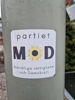 Sticker for the political party Månskliga rättigheter och Demokrati (Human Rights and Democracy) in Sweden.  The sticker features the party's logo, which includes the letters "MOD" and a stylized sunflower.