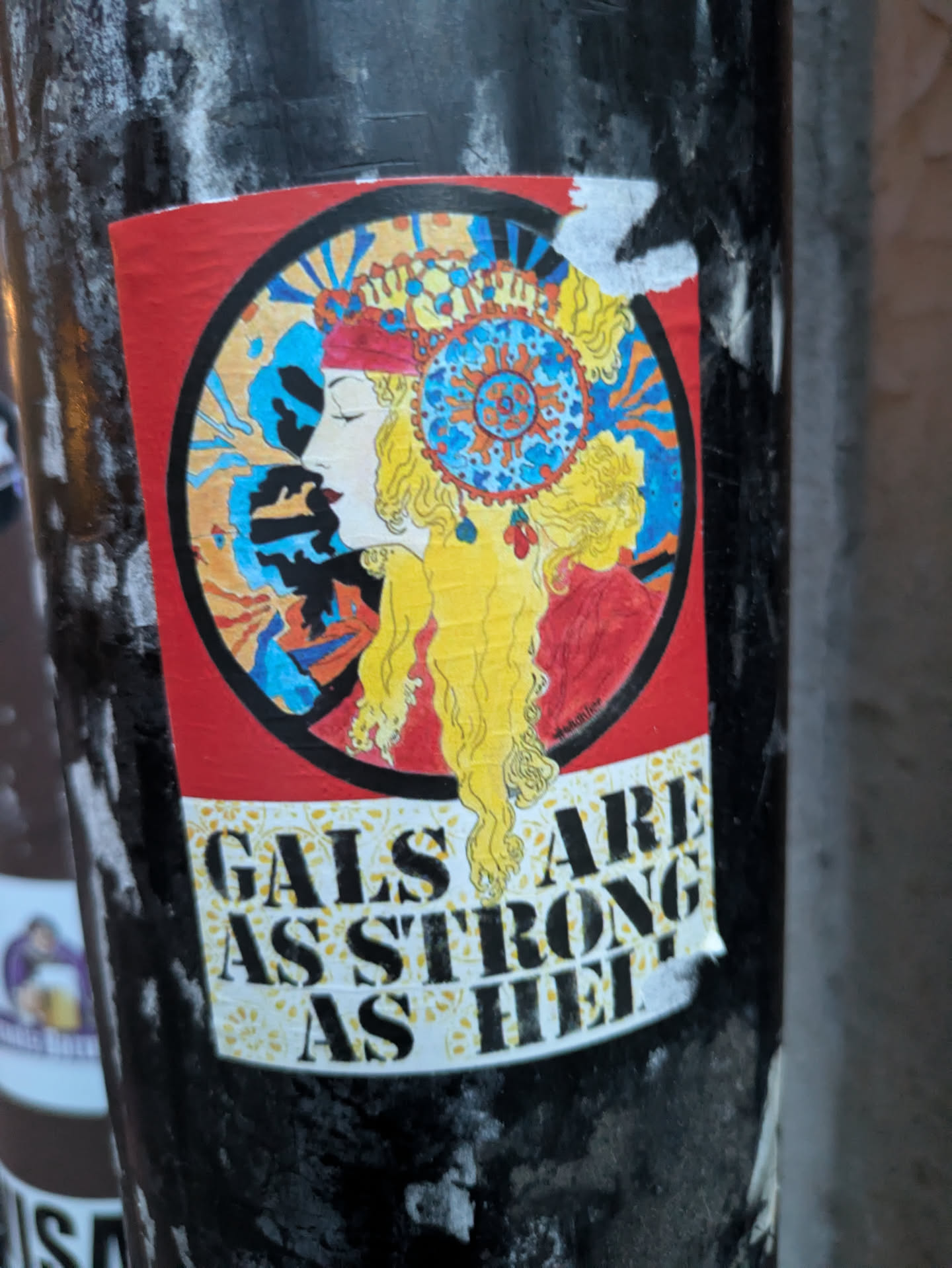 The sticker features a circular illustration of a woman's profile with long, flowing blonde hair. She is adorned with an elaborate, colorful headdress featuring geometric patterns and beads in blue, yellow, and red. Her profile is serene, with eyes closed and red lips. The background within the circle is a vibrant mix of blue, orange, and black abstract shapes, possibly representing rays or energy. Below the circular artwork, the text 'GALS ARE STRONG AS HELL' is displayed in bold, black, sans-serif lettering against a white background with a subtle yellow floral pattern. The sticker has a slightly worn appearance with some peeling at the edges, suggesting it has been applied to a surface.