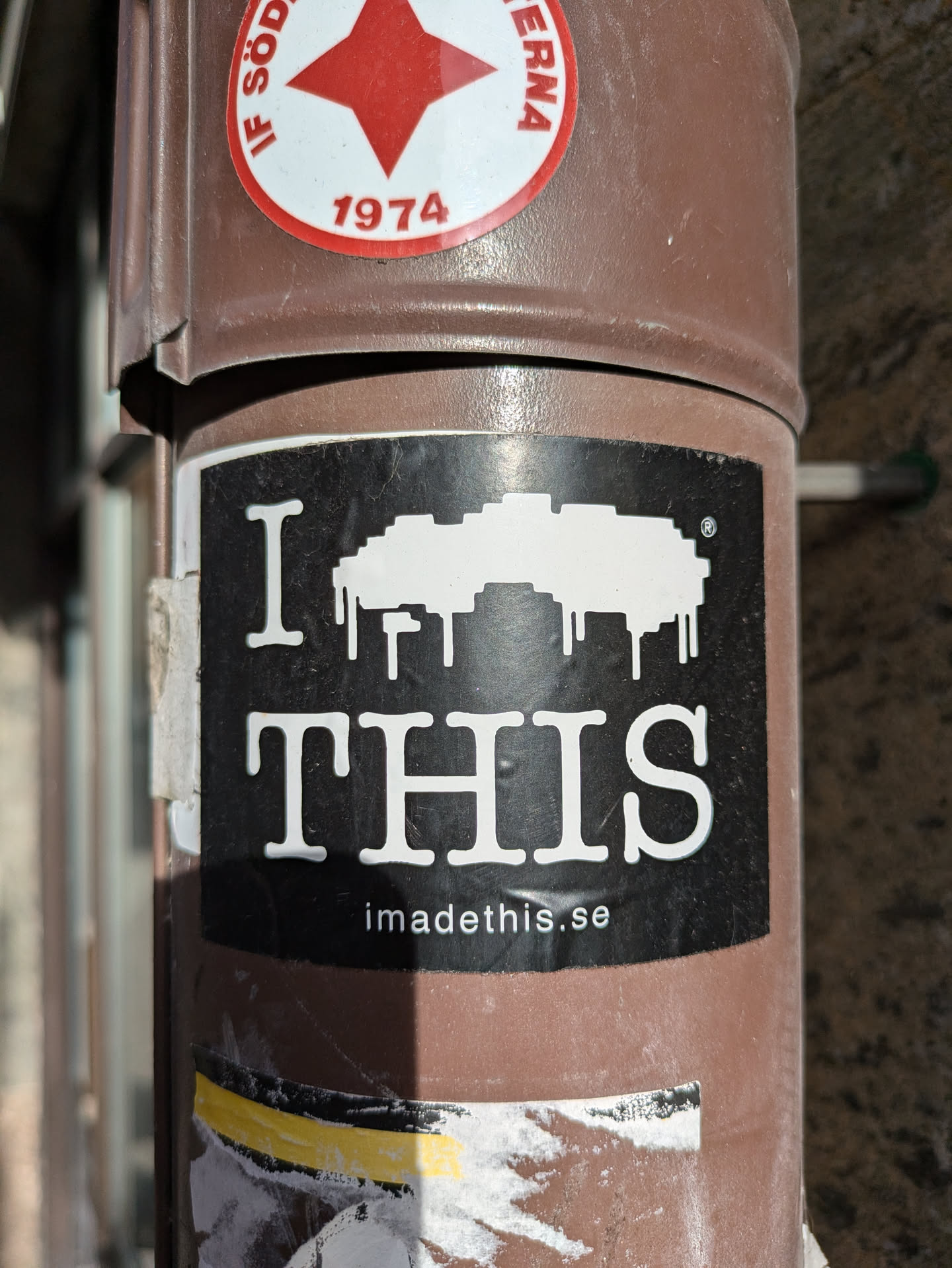 Black and white sticker with a pixel art design and the text "I \u2764 THIS" and a website address at the bottom. The sticker is affixed to a brown pole.