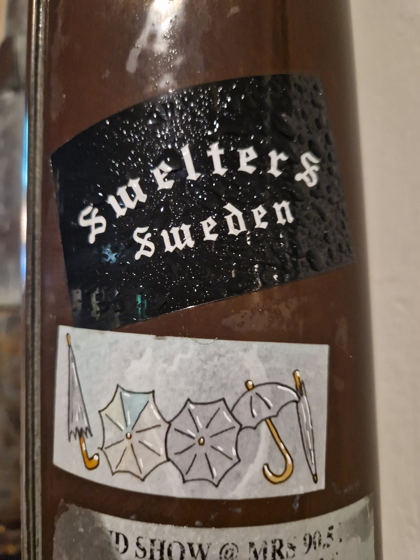A sticker featuring the text Swelters Sweden on a black background, and another sticker with three umbrellas on a brown background. There's also some text partially visible at the bottom that seems to mention a show and a location (MRS 905).