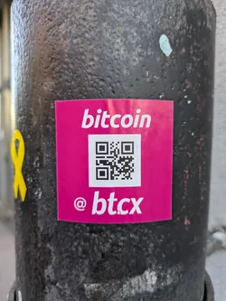 A pink square sticker with the word "bitcoin" at the top, a QR code in the middle, and "@btc.x" at the bottom. It is affixed to a dark gray or black cylindrical object.