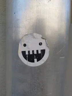 A round sticker with a minimalist design of a face. It has two small dots for eyes and a simple black and white design for a mouth. The sticker is affixed to a gray metal pole.