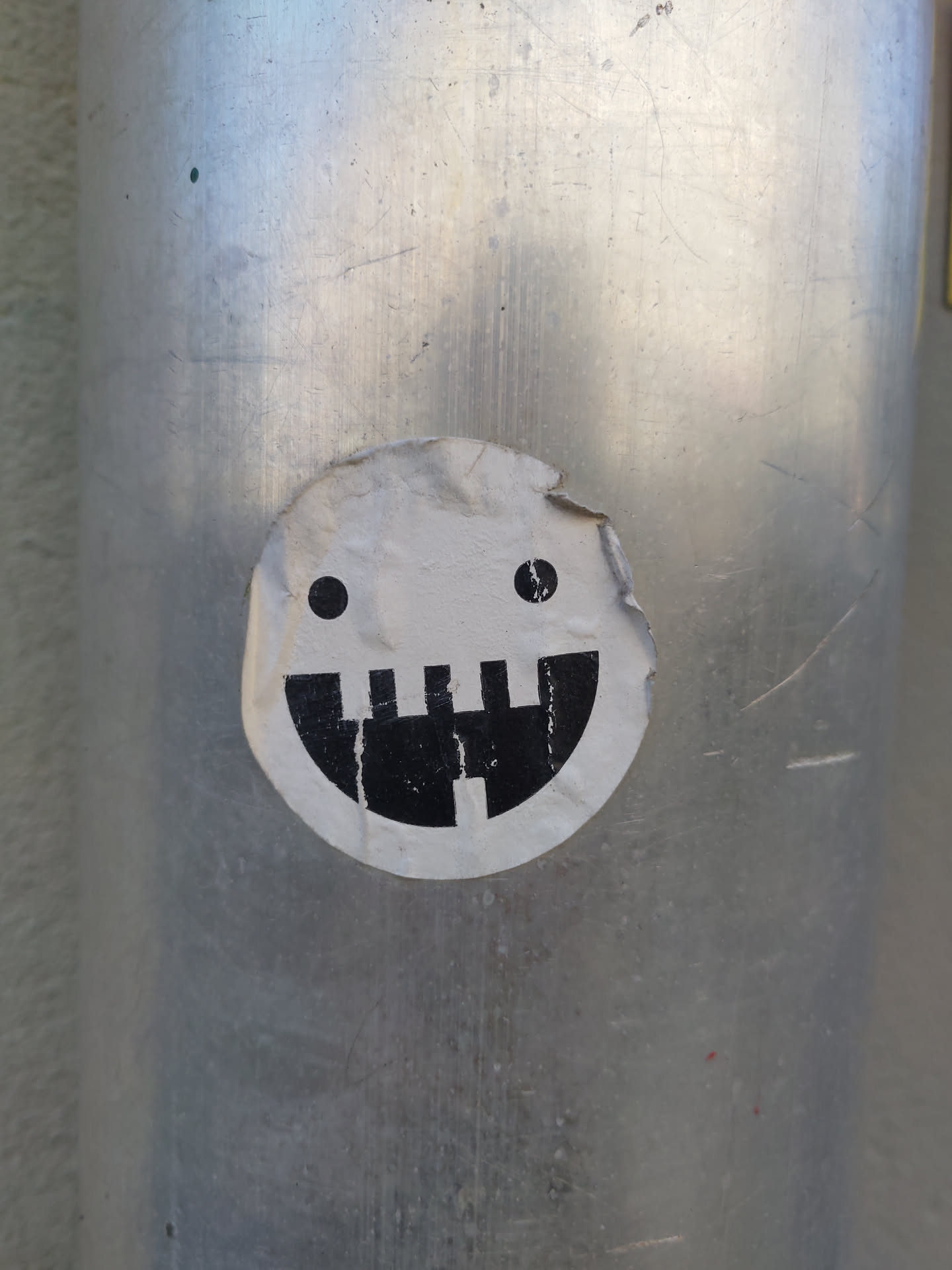 A round sticker with a minimalist design of a face. It has two small dots for eyes and a simple black and white design for a mouth. The sticker is affixed to a gray metal pole.