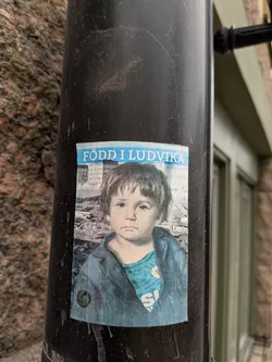 The sticker features a close-up portrait of a young child with striking blue eyes and tousled brown hair. The child is wearing a blue jacket over a turquoise shirt with white lettering. The background of the sticker appears to be a faded image of buildings. The top of the sticker has a blue banner with white text. The overall impression is one of a vintage or nostalgic photograph.