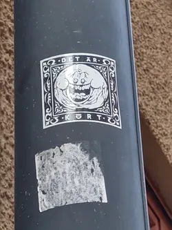 Black and white sticker with a graphic of a smiling, somewhat unsettling, round face inside an ornate frame. The text ".DETÄR. KØRT." is visible above and below the image.