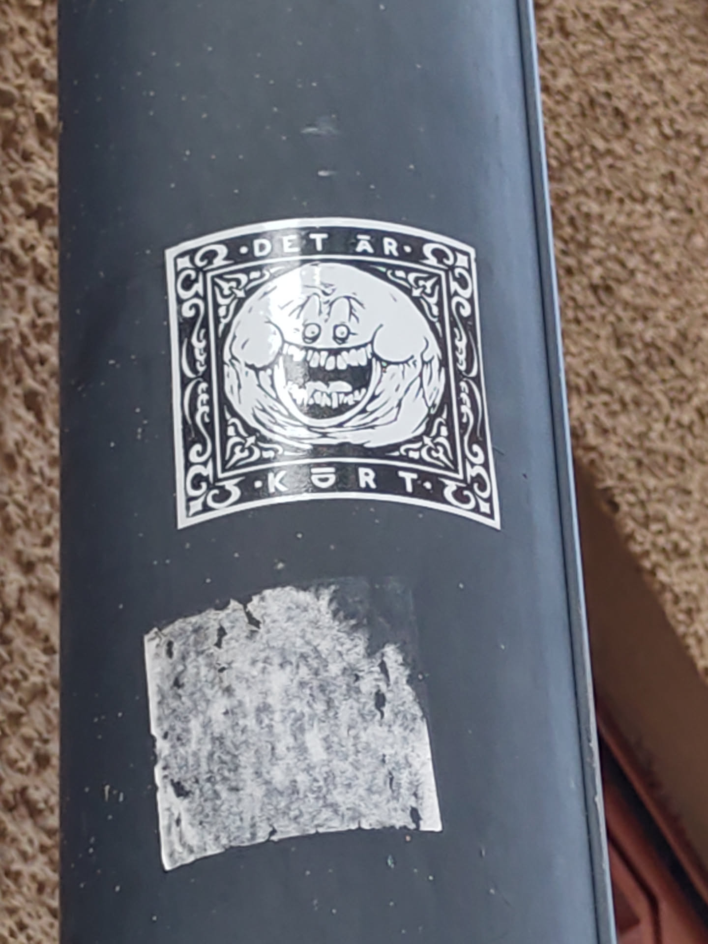 Black and white sticker with a graphic of a smiling, somewhat unsettling, round face inside an ornate frame. The text ".DETÄR. KØRT." is visible above and below the image.