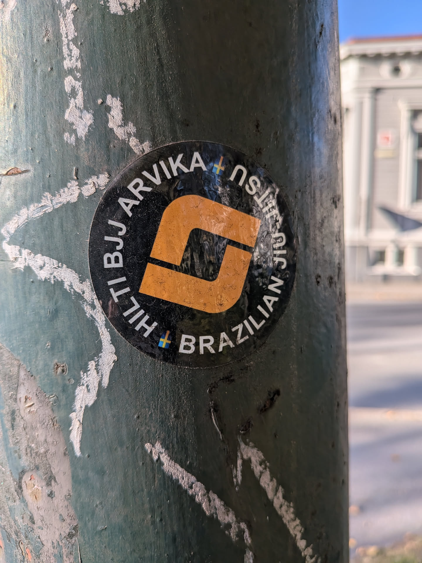 The sticker is circular with a black background. In the center, there is a stylized orange 'J' shape, which is a common symbol in Jiu-Jitsu. The text 'TI BJJ ARVIKA' is written in white capital letters in an arc above the 'J', and 'BRAZILIAN' is written in white capital letters in an arc below it. Small rainbow-colored crosses are visible between some of the letters, and a Swedish flag symbol is also present. The sticker appears slightly worn and is affixed to a dark, weathered pole.