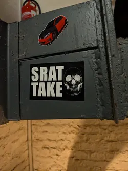 Street sticker A rectangular black sticker affixed to a dark grey metal structure. On the left side, the text 'SRAT' is stacked above 'TAKE' in bold, white, condensed sans-serif capital letters. To the right of the text is a high-contrast white illustration of a front-facing human skull.