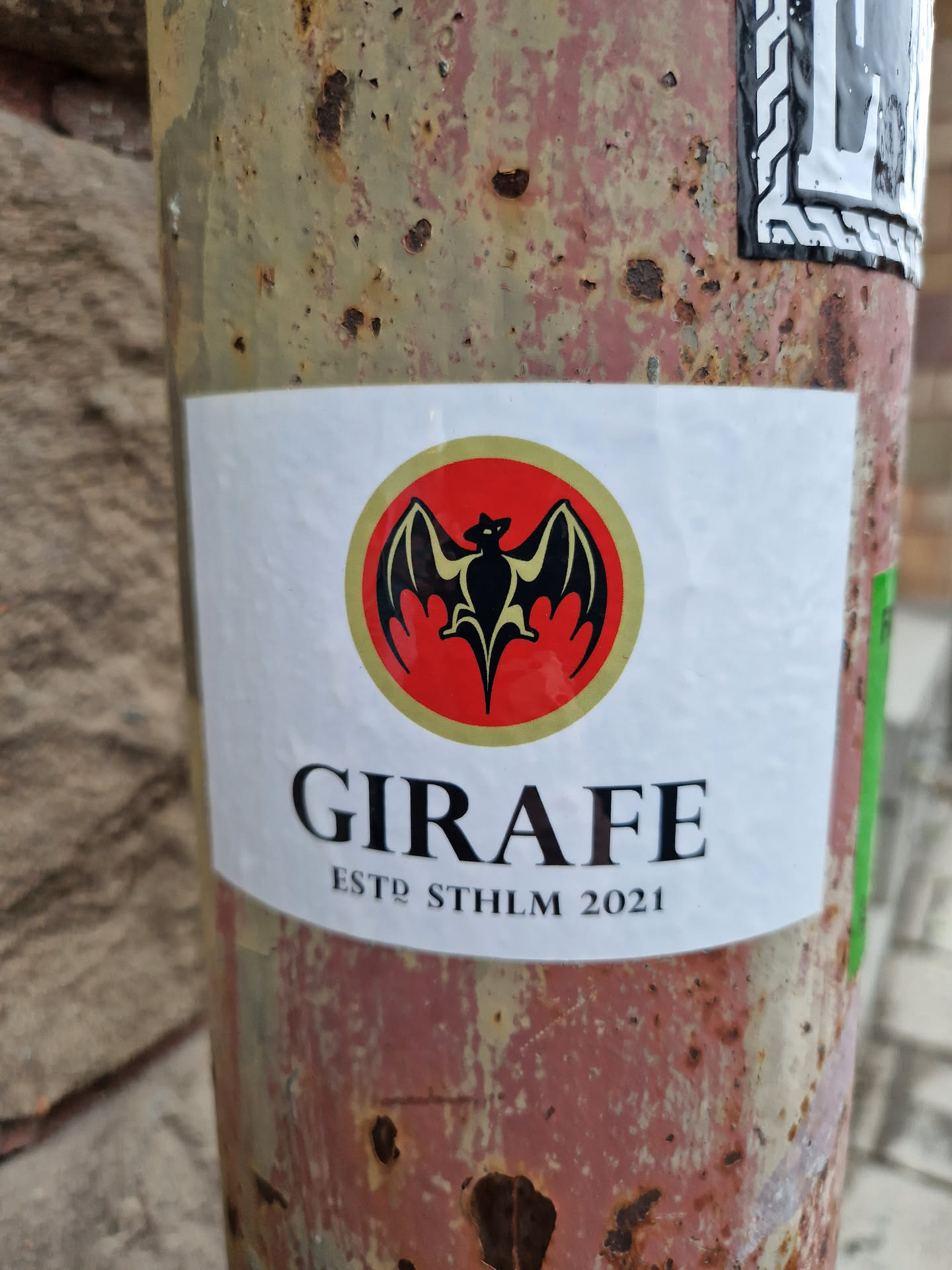 A sticker with a bat logo inside a red circle and the text GIRAFE ESTD STHLM 2021. The sticker is affixed to a rusty metal pole.