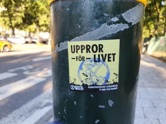 A yellow sticker featuring the text "Uppror - för - livet" (Rebellion for life) along with an illustration of the earth with a cross over it and small plants. The sticker also mentions climate protests in Stockholm from 17th August onwards. It has the Extinction Rebellion logo on it.
