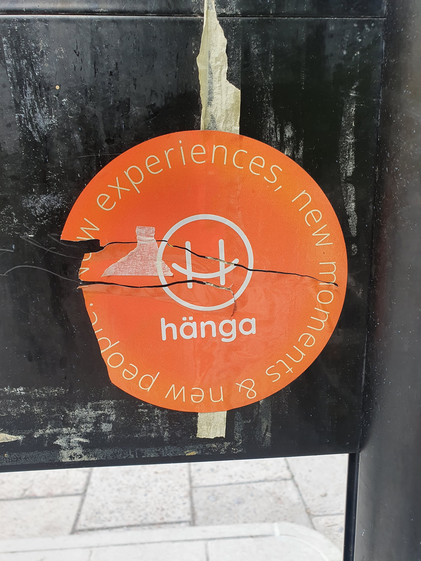 An orange round sticker with the word "hänga" in the center and the phrase "New experiences, new moments & new people" written around it. The sticker is slightly damaged and torn.