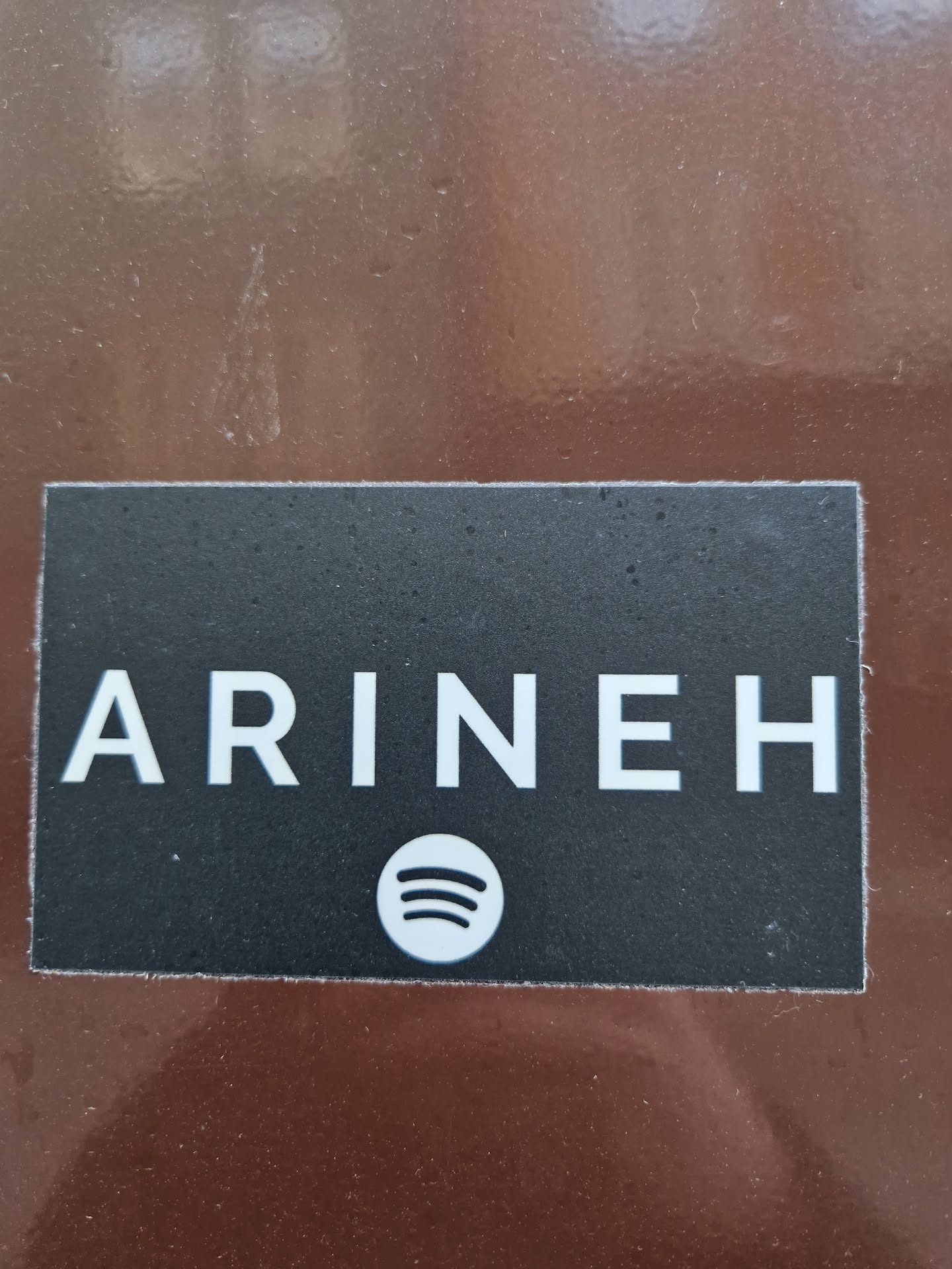 A rectangular black sticker with the name ARINEH written in white capital letters and the Spotify logo below it. The sticker is on a brown surface.