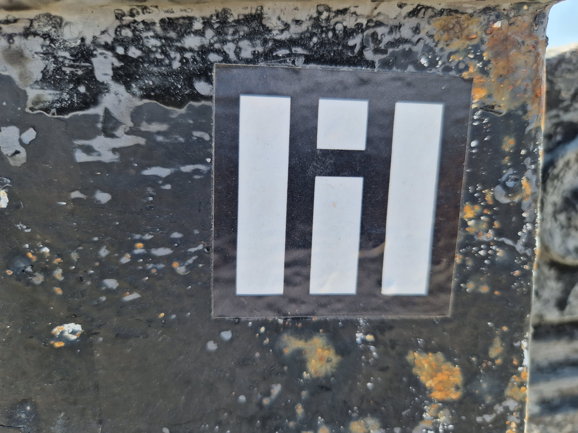 A square black and white sticker with a stylized 'Hi' or similar abstract design. The sticker is affixed to a dark, textured, possibly metallic surface with signs of wear and rust.