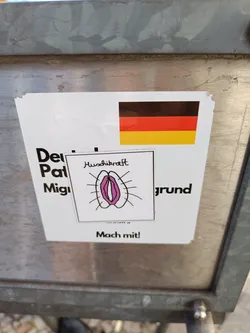 A sticker featuring a simple drawing of a vulva, the German flag, and text that appears to be cut off.  The visible text includes the word 'Mach mit!' which translates to 'Join in!' in English.
