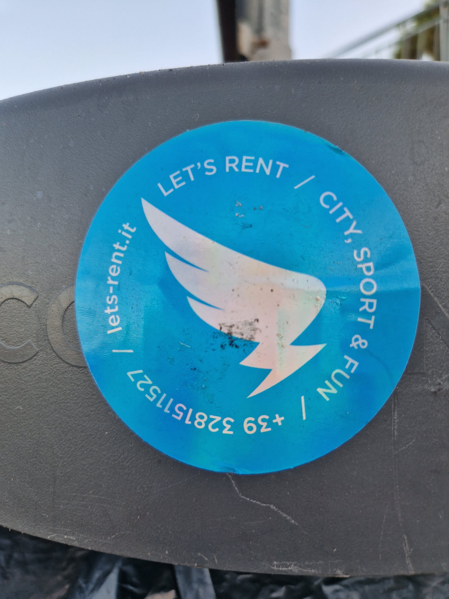 A round sticker with a blue background featuring a stylized wing design and the text "LET'S RENT | CITY, SPORT & FUN | +39 328 1515231 | lets-rent.it".