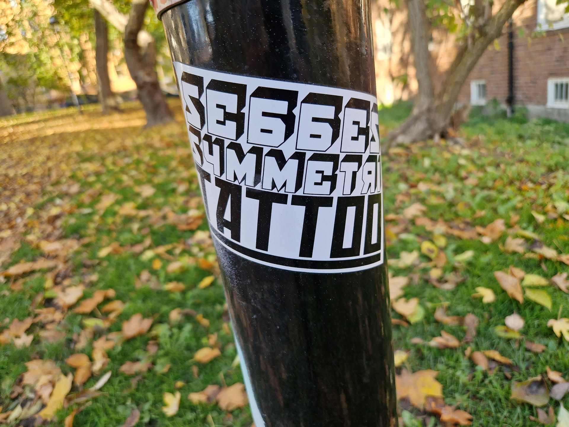 A black and white sticker with the text "SE66EX Symmetrical Tattoo" in a stylized font. The sticker is affixed to a black pole in an outdoor setting.