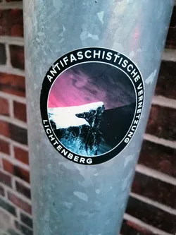 A round sticker with a dark background and a pink and purple sky. The text around the circle says "Antifaschistische Vernetzung Lichtenberg".