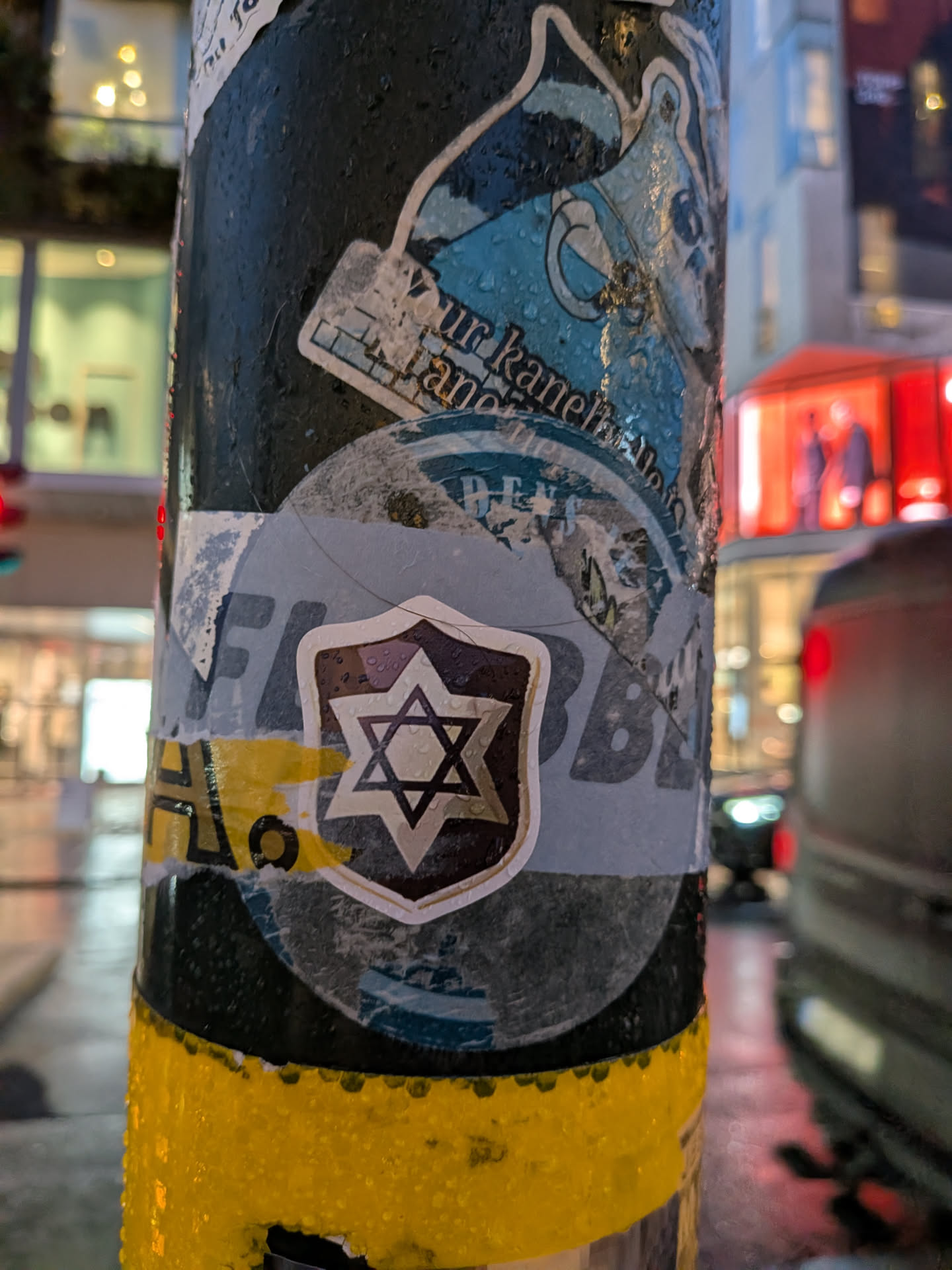 The central sticker features a stylized Star of David within a shield-like emblem. The star is black with a white outline, set against a dark background with a textured, almost metallic appearance. The shield itself has a dark brown or black border and a lighter, greyish interior. The sticker is layered over other, partially visible stickers, one of which has a blue and white design with some text that is difficult to read. The entire pole and stickers appear wet, with visible water droplets, suggesting it might be raining or has recently rained. The background is blurred, showing urban elements like buildings and lights.