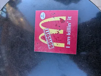 The sticker is rectangular with a vibrant red background. It prominently displays the iconic golden arches (McDonald's 'M' logo) in yellow. Overlaid on the yellow arches and the red background are numerous dark red splatters, resembling blood. A white rectangular banner with a red border is angled diagonally across the 'M', bearing the word "BOYCOTT" in bold red capital letters. To the right of the 'M', in white sans-serif font, is the phrase "I am killin' it". In the top-left corner, there is a small white circular element containing an Instagram logo and the handle "@EPYUS" in black text. The sticker appears slightly worn with some dust and minor scratches.