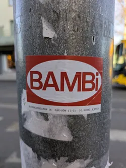 This sticker features the word 'BAMBI' in bold, white letters against a red, rounded rectangular background. Below the logo are details including an address and opening hours. The sticker is affixed to a gray metal pole.