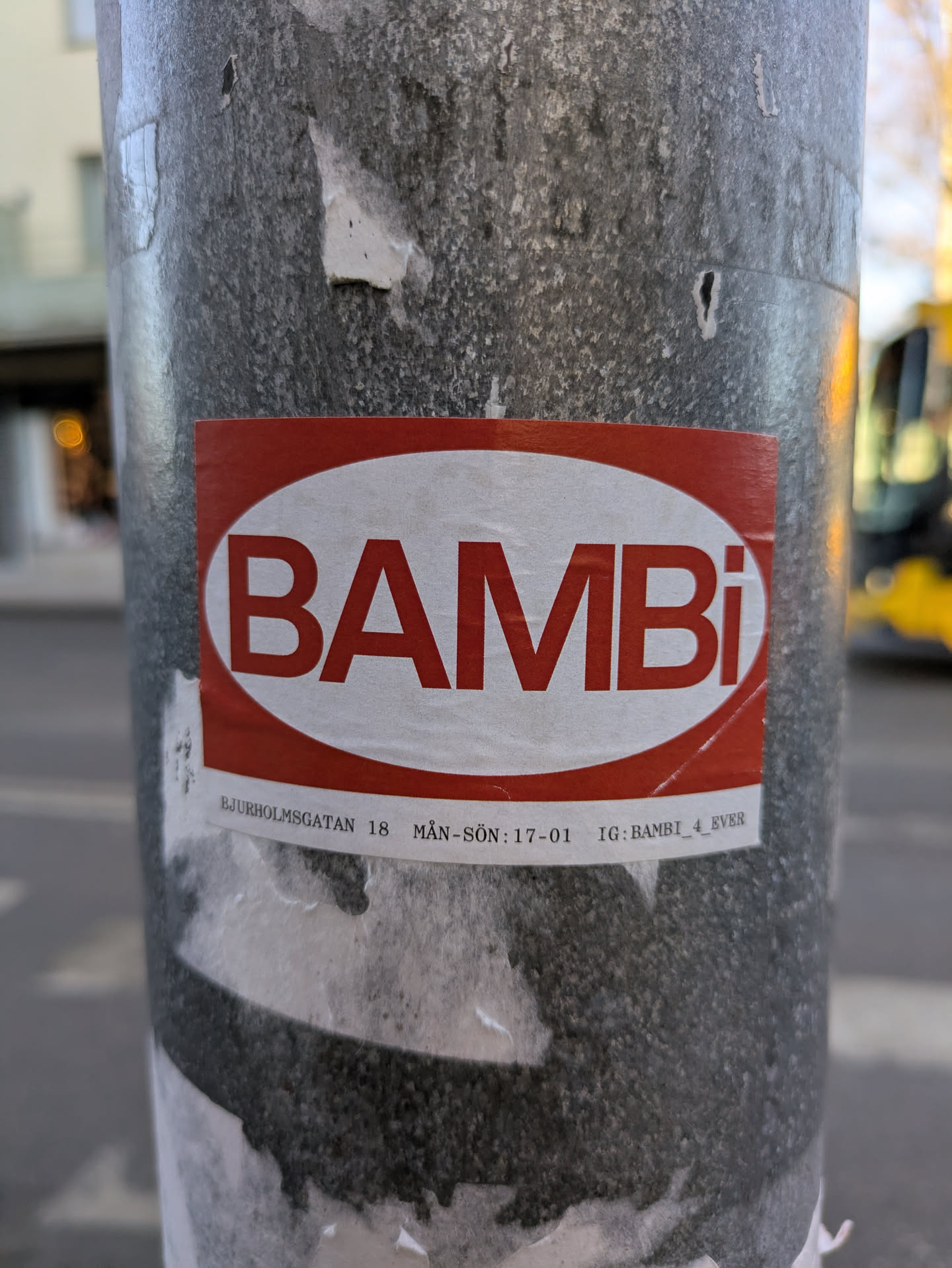This sticker features the word 'BAMBI' in bold, white letters against a red, rounded rectangular background. Below the logo are details including an address and opening hours. The sticker is affixed to a gray metal pole.