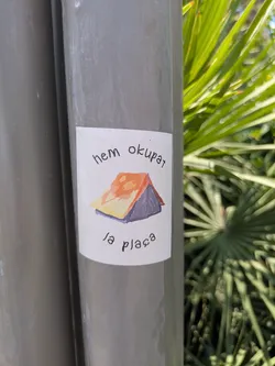A sticker on a pole features a simple drawing of a tent with an orange roof and purple base. The text 'Hem Okupat la plaça' is written above and below the image of the tent. The background is a leafy plant.