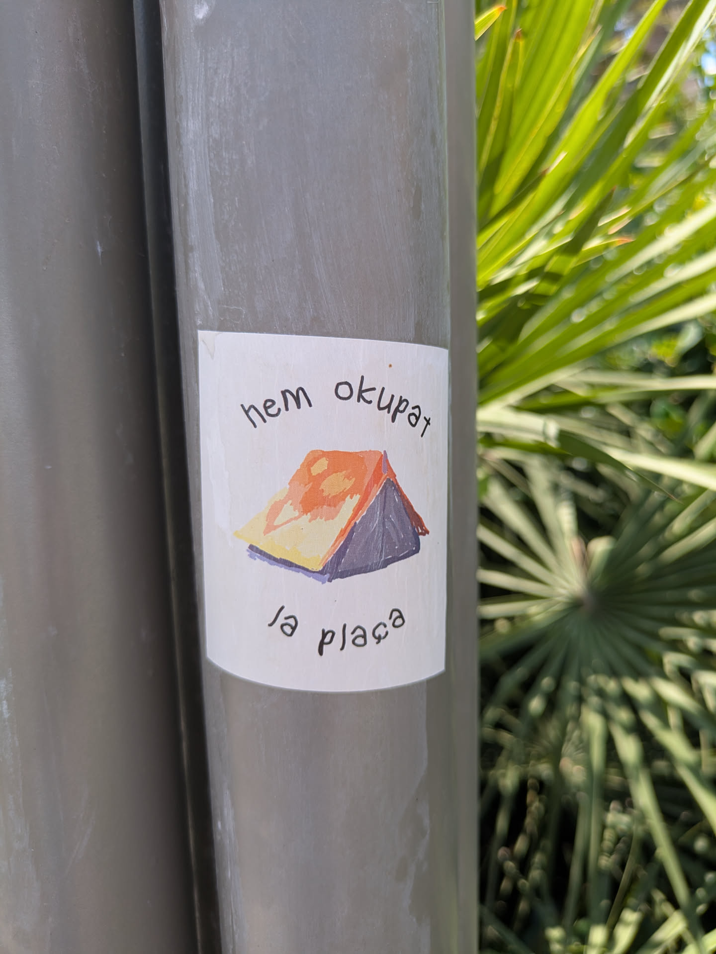 A sticker on a pole features a simple drawing of a tent with an orange roof and purple base. The text 'Hem Okupat la plaça' is written above and below the image of the tent. The background is a leafy plant.
