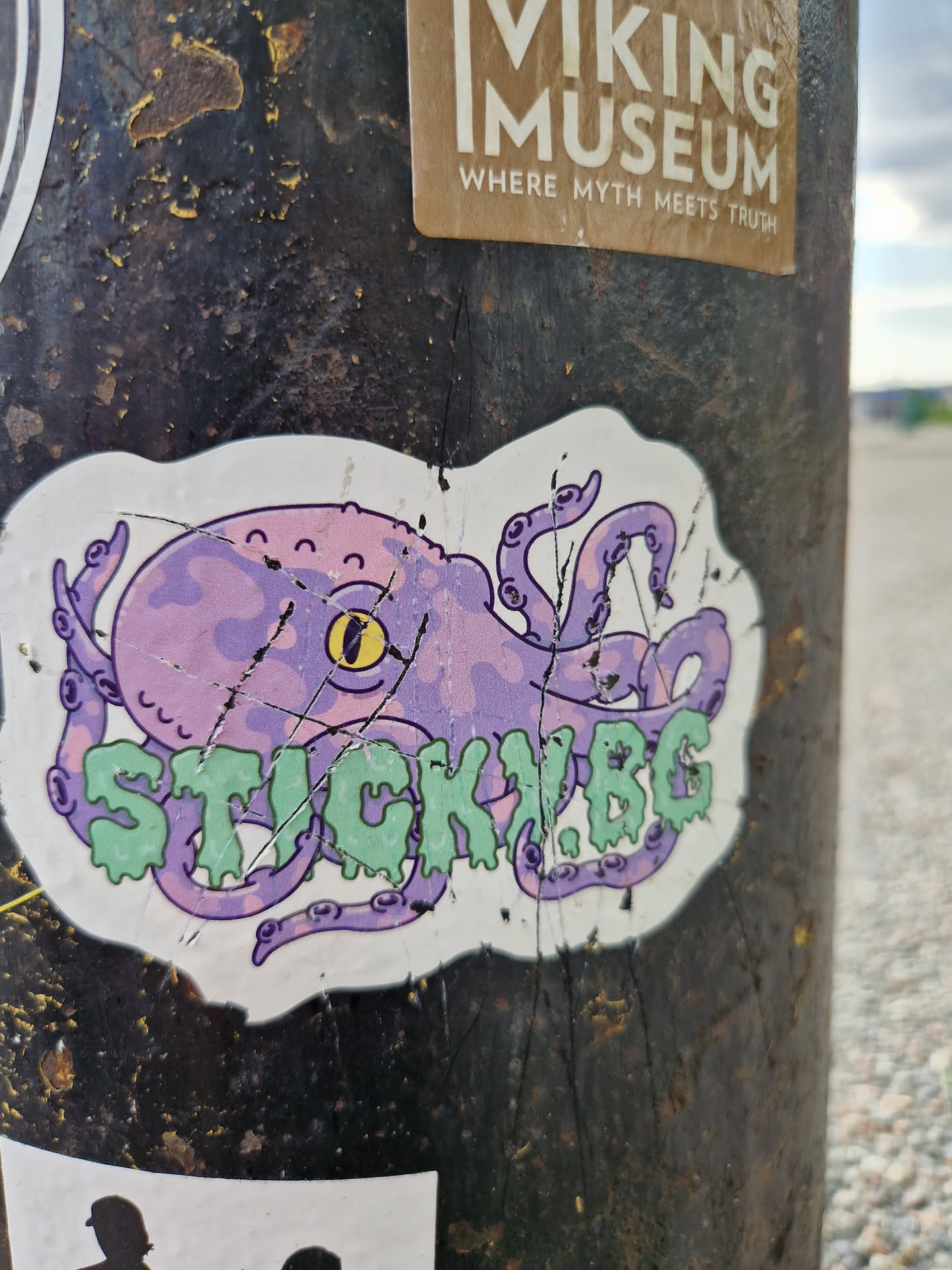 A sticker featuring a cartoon octopus in purple and light green colors. The octopus has a friendly expression and its tentacles surround the text "STICKY.BG". The sticker appears to be slightly damaged, showing some tears and scratches.