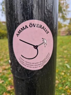 Pink round sticker with a graphic of a breast with a line shooting out of the nipple. The text around says Amma Överallt (Grandma Everywhere) and lists places where it can be found in Swedish.
