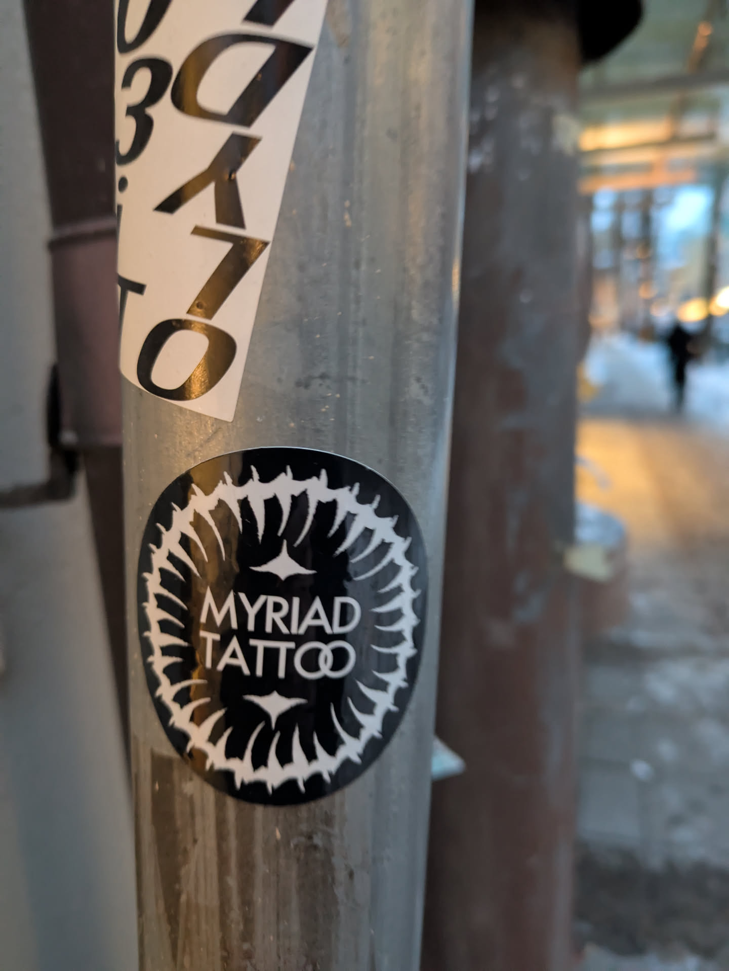 Black and white sticker with a circular design and the text 'MYRIAD TATTOO' in the center. The circular design looks like a stylized flower or mandala. The sticker is affixed to a metal pole.