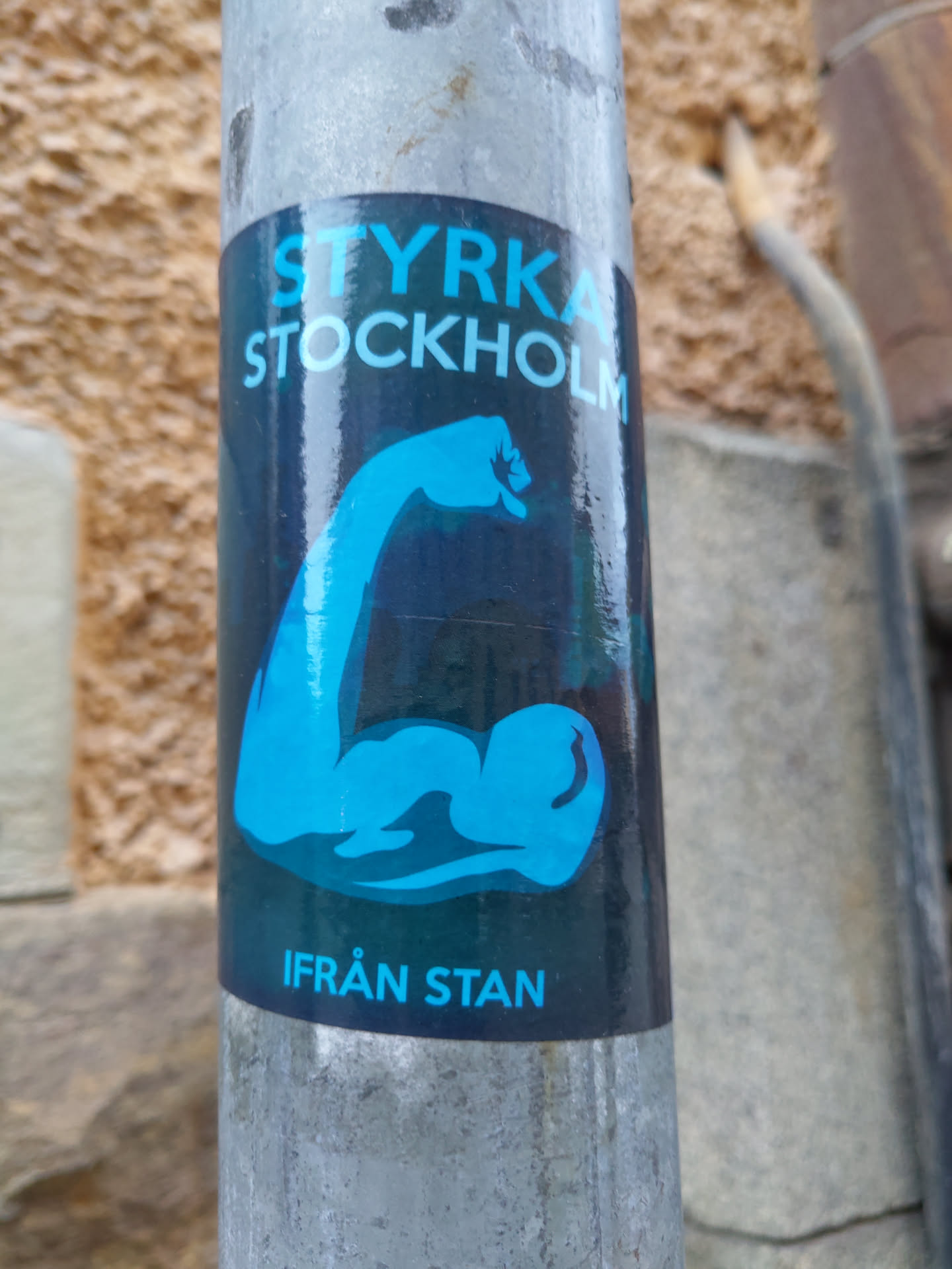 A sticker featuring a bicep flex graphic with the text "Styrka Stockholm" (Strength Stockholm) and "Från Stan" (From Stan) in light blue on a dark background. The sticker is applied to a metal pole.