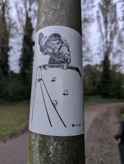 A black and white illustration sticker featuring a person playing billiards. The sticker is applied to a pole outdoors.  The style is simple line drawing. A small logo is visible in the bottom right corner.
