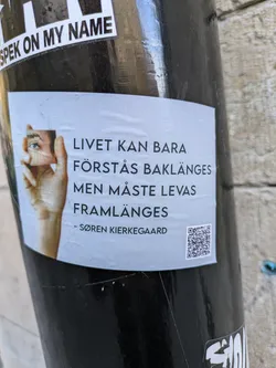 A sticker featuring a quote by Søren Kierkegaard. The quote in Swedish translates to "Life can only be understood backwards; but it must be lived forwards."  The sticker also includes an image of an eye and a QR code.
