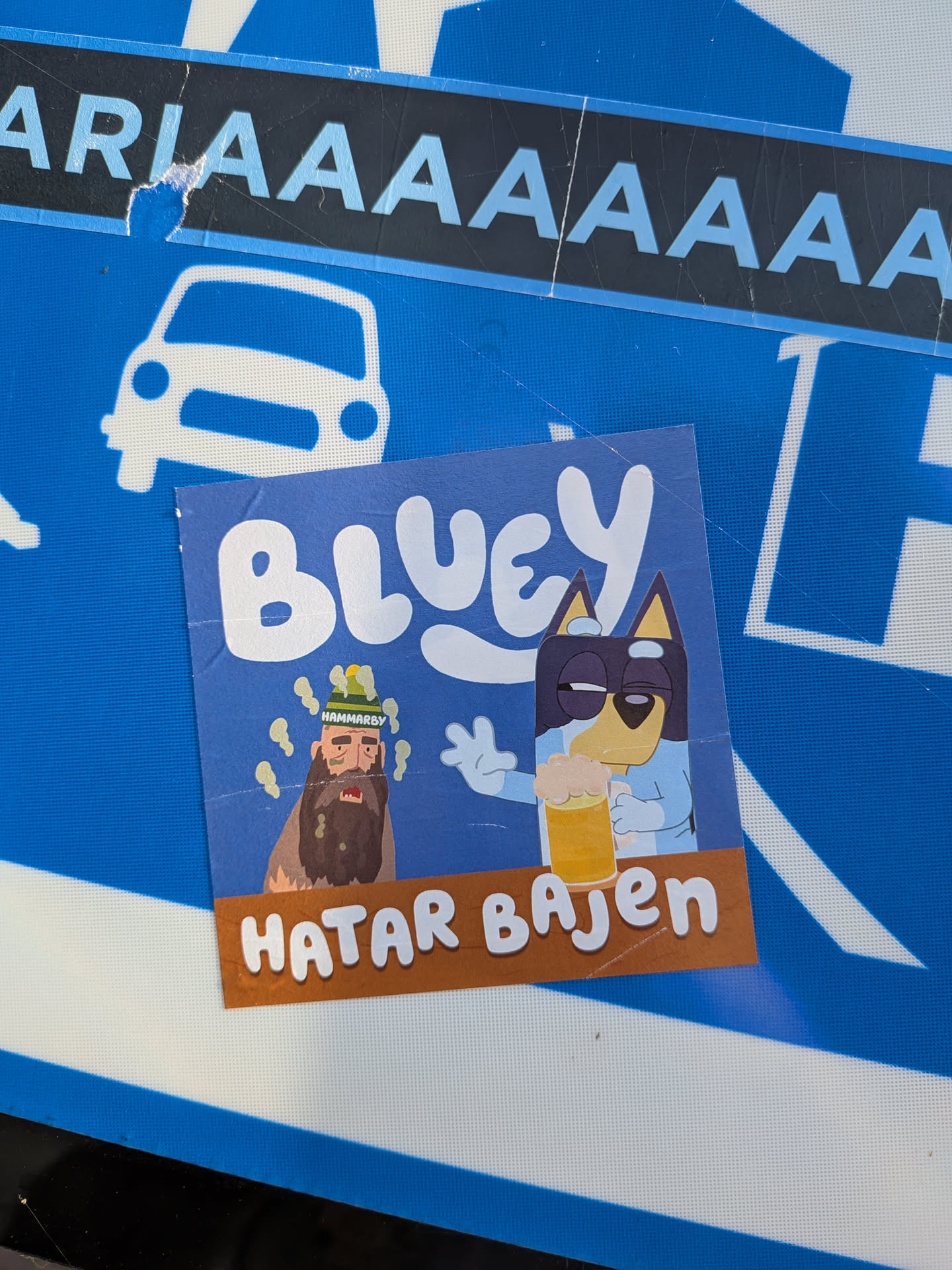 The sticker is blue and features characters from the show 'Bluey'. In the center, Bluey character Bingo is depicted holding a pint of beer, looking relaxed with sunglasses on. To the left of Bingo is a character with a beard and a green hat that says 'HAMMARBY'. There are yellow, wavy lines emanating from this character, possibly representing an idea or a feeling. Below the characters is a brown banner with white text.