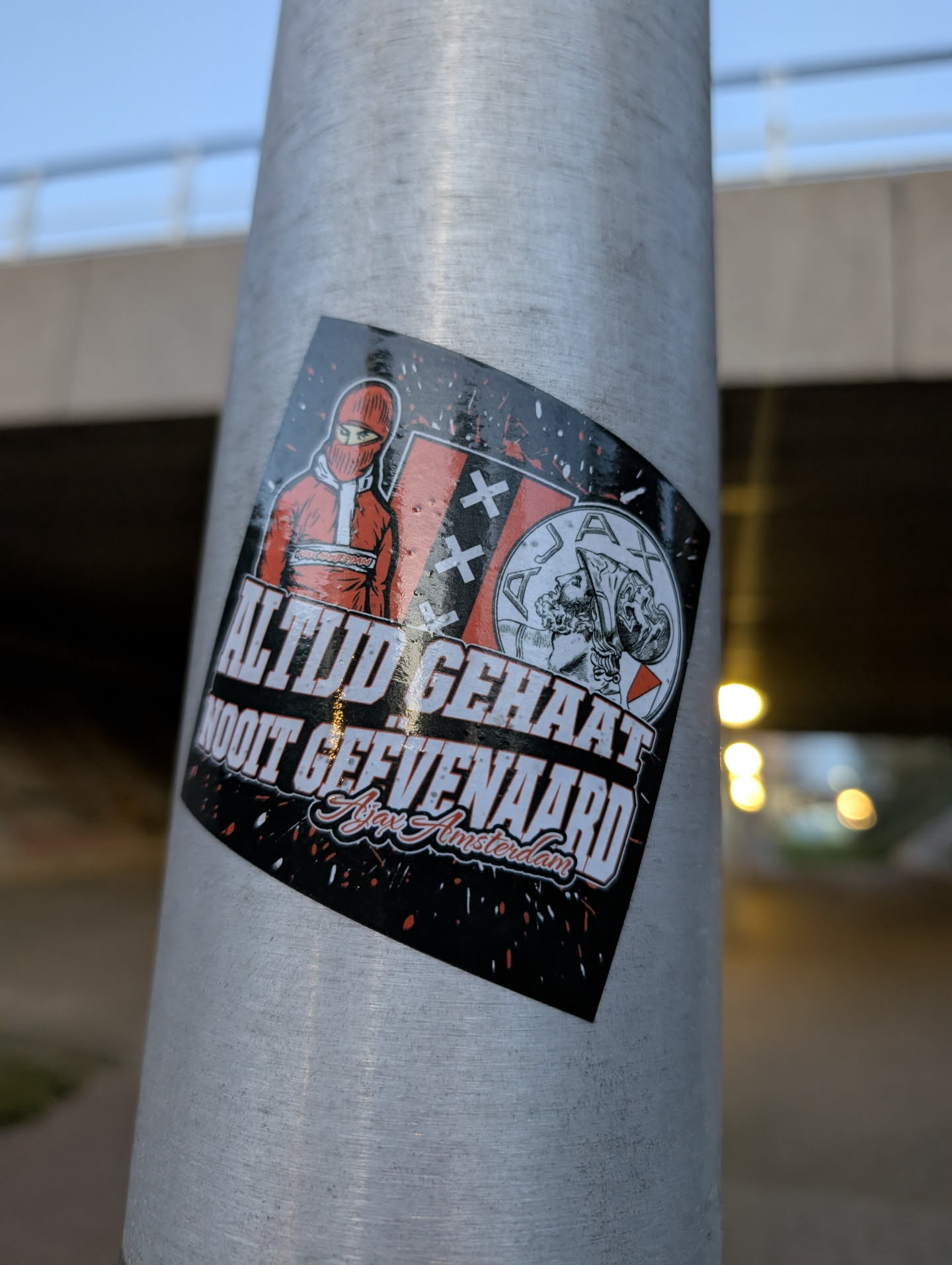 The sticker is predominantly black with red and white accents. On the left, it features a stylized illustration of a person wearing a red balaclava and a red jacket with white trim. To the right of this figure, there is a representation of the Ajax Amsterdam crest, which includes three white crosses and a depiction of Ajax himself. Below these images, bold white text reads 'ALTIJD GEHAAT' and 'NOOIT GEEVEE NAARD', with 'Ajax Amsterdam' written in a stylized script below. The background of the sticker has a distressed, splattered effect in red and white.