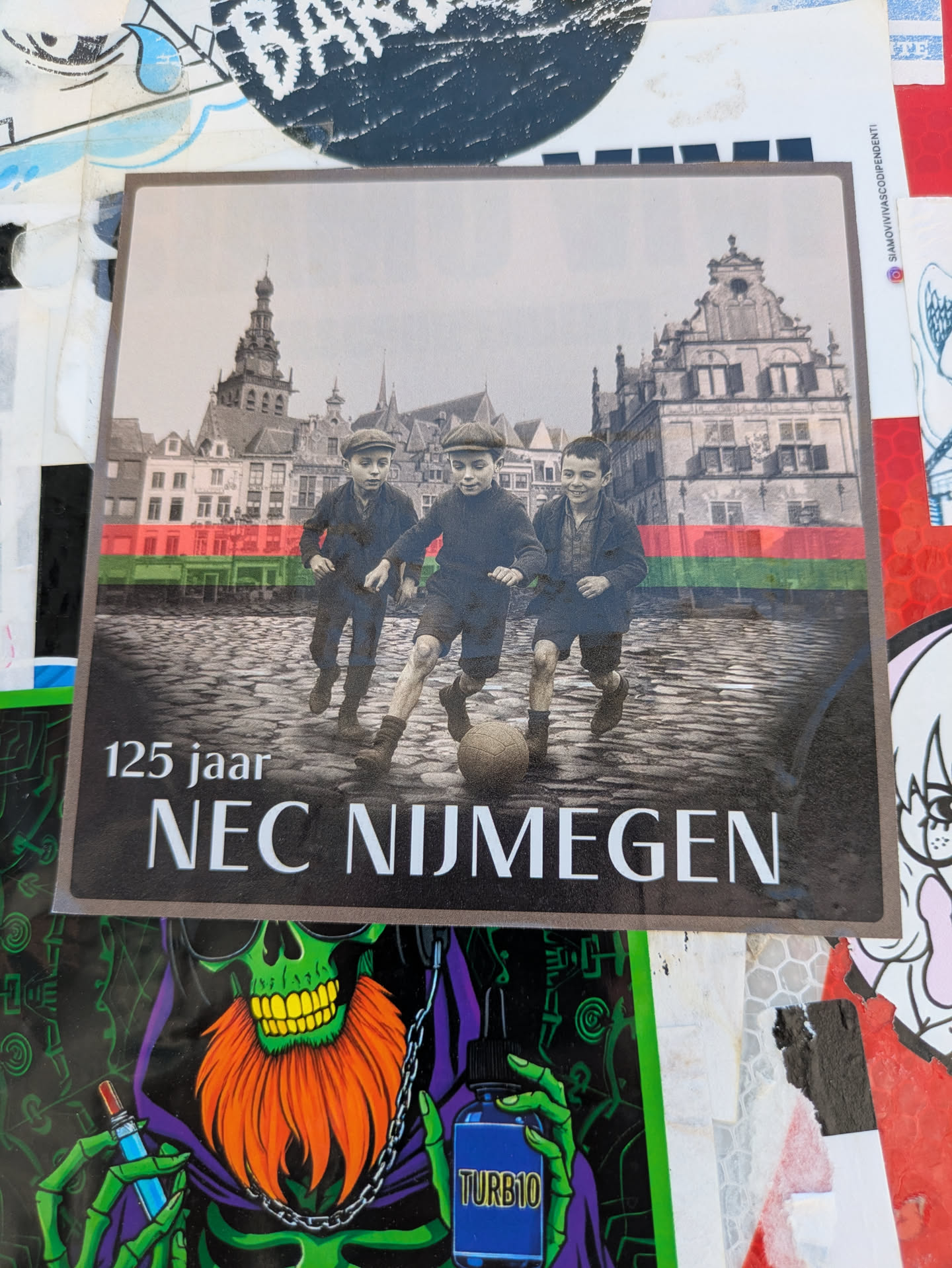 This sticker features a sepia-toned image of three children playing football in front of a historic European city. A red, black, and green horizontal line is featured on the middle of the image. The sticker commemorates '125 jaar NEC NIJMEGEN', indicating an anniversary celebration for a sports club. The vintage photography and classic typography give the sticker a nostalgic and celebratory feel.