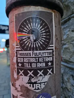 A black and white sticker featuring a stylized image of Idi Amin with syringes radiating outwards. A rainbow prism is shown emanating from his eye.  The text in Swedish translates to "Frasses' full move gives astral ketamine to Idi Amin."