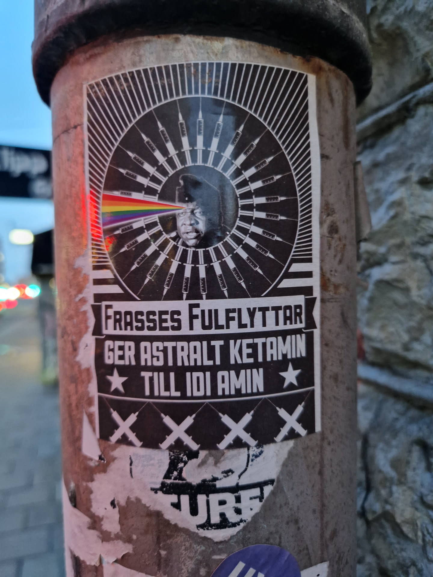 A black and white sticker featuring a stylized image of Idi Amin with syringes radiating outwards. A rainbow prism is shown emanating from his eye.  The text in Swedish translates to "Frasses' full move gives astral ketamine to Idi Amin."