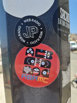 A red circular sticker features an abstract design composed of black shapes, blue eyes, and geometric patterns.  At the bottom, the text "ABAMC" is visible. A small QR code is also included within the design.